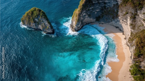A beach is seen surrounded by high cliffs and large rock formations. Waves crash on the shore. The sun shines brightly on the blue water. This scene shows a tropical coastal area.