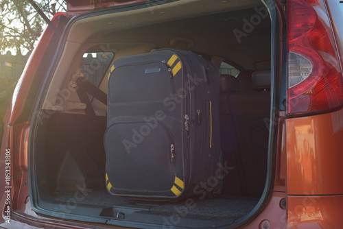 Large black suitcase with yellow accents standing upright inside the trunk of an orange car, ready for a trip.