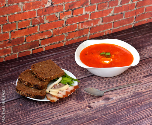 A plate of hot freshly cooked Slavic soup Borscht with sour cream, next to it a plate with rye bread, lard, garlic and green onions.