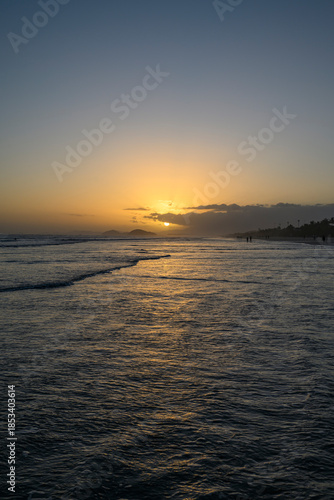 Praia ao pôr do sol com reflexos dourados na areia molhada, ondas suaves e silhuetas humanas. Cena tranquila e inspiradora de beleza natural e contemplação.