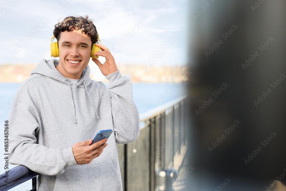 Fototapeta premium Young man in grey hoodie with headphones and mobile phone on embankment