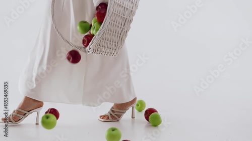 A woman with a basket of apples walks in shoes on a white background. Apples fall to the floor.