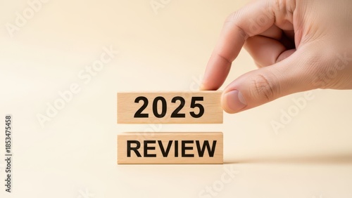 A hand places a wooden block with '2025' on top of another block labeled 'REVIEW'. Represents year-end analysis, performance review, and future assessment.