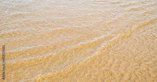 Fotografie Clear shallow sea water with gentle waves over golden sand, warm coastal texture in horizontal format, natural beach background perfect for summer, travel or vacation designs