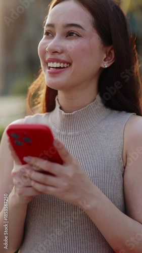 Portrait of happy Asian woman flirts with boyfriend online via smartphone in city vertical shot. Brunette Chinese lady smiles reading message in chat on mobile phone on town street
