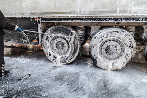 Man is washing a truck tire with a hose. The tire is covered in foam and the man is using a brush to scrub it
