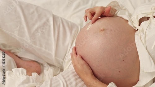 Pregnant woman lies on bed and applies cream on belly. Female in last month of pregnancy uses anti stretch mark cream from jar. She takes product with finger and gently massages abdomen. Body care rou