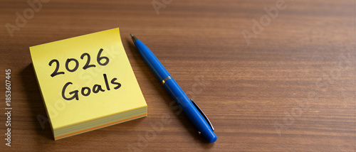 2026 Goals handwritten on a yellow sticky note next to a blue pen on a wooden desk. New Year's Resolution.
