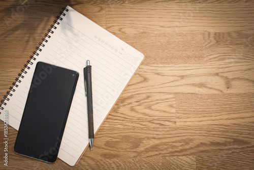 Top view of blank notebook with pen and smartphone on wooden desk with copy space.