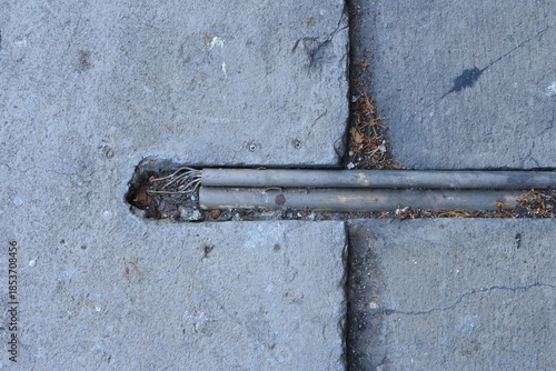 High angle view of two weathered plastic conduits embedded in a gap between uneven concrete slabs. On the left side, a section of one conduit is damaged, revealing a bundle of insulated wires.
