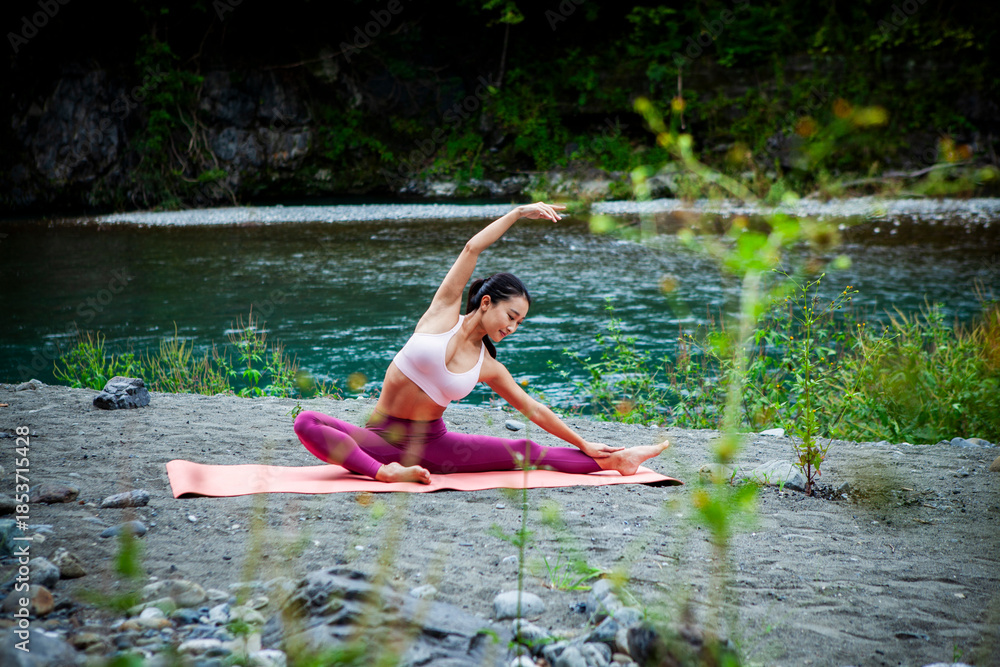 Fototapeta premium Young Japanese woman practicing side stretch yoga pose by a river, active outdoor wellness lifestyle with mindfulness, sustainability and mental wellbeing focus