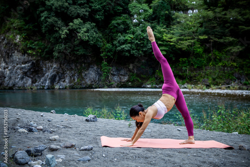 Asian woman practicing advanced yoga balance pose outdoors by a river, strength and mental wellness lifestyle in nature