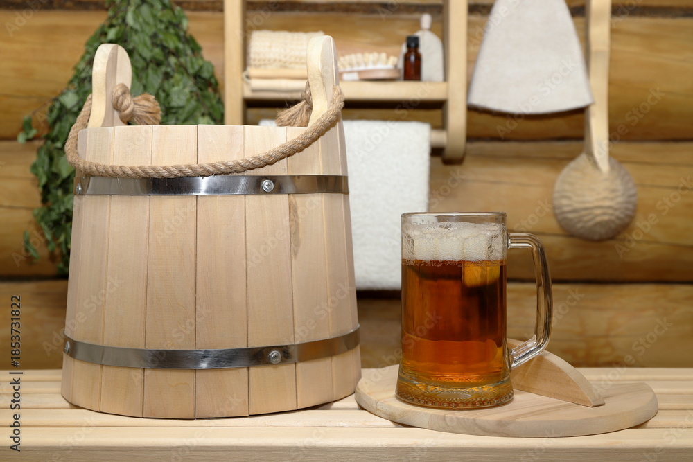 Φωτογραφία A glass mug of light beer and wooden bucket for bath treatments stand in the sauna interior on a bench against log wall with birch broom, ladle, hat and shelf with bath accessories and a white towel