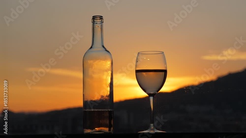 Rick natural colour of sunset afterglow, transparent bottle and glass of light red wine standing on table against window. Still life shot with blurred orange sky on background