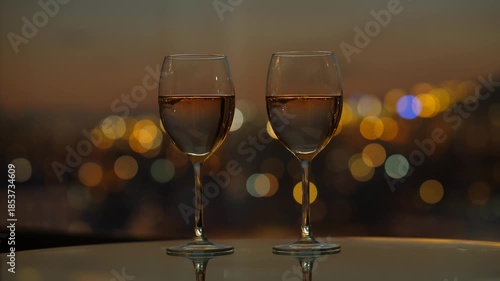 Two glasses with white wine against blurred city lights on background, time lapse shot of nightfall, sky quickly become dark. Neat colours of large round twinkling lights, tender natural light