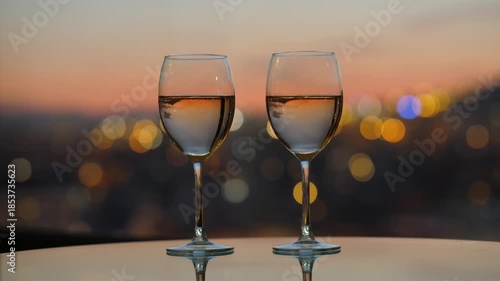 Wineglasses for two standing at glass table, blurred city lights and neat darkening sky on background, dusk time after sunset.