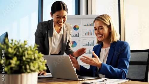 Two businesswomen collaborating on laptop in modern office setting with graphs