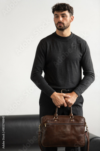 Professional male model in charcoal crewneck knitwear holding a dark brown leather briefcase in a minimalist studio setting