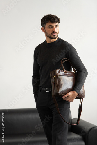 Professional male model in charcoal crewneck knitwear holding a dark brown leather briefcase in a minimalist studio setting