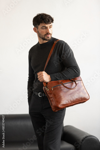 Professional male model in charcoal crewneck knitwear holding a light brown leather briefcase in a minimalist studio setting