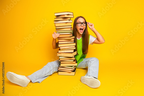 Schilderij op canvas Young woman student with a tall stack of book smiling against a bright yellow ba