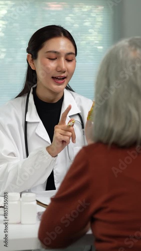 Young Asian physician in a white coat explaining the dosage of medication from a pill bottle to an elderly woman during a medical consultation in a modern hospital clinic room