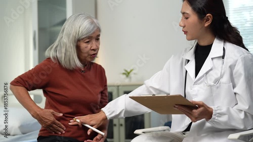 Elderly asian woman describing her stomach ache symptoms to a female doctor during a medical checkup in a hospital examination room, showing the painful area for diagnosis
