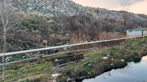 A river in the forest is covered with a thick layer of trash; a terrible smell and view; a mountain of trash behind. Underneath are pipes collecting biogas methane.