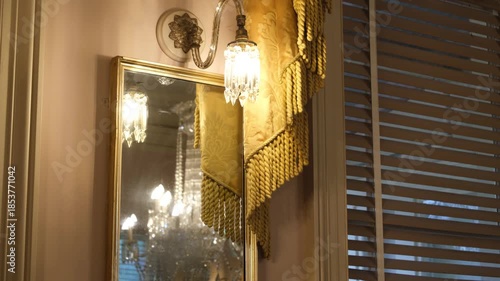 An elegant vintage wall sconce with a crystal glass shade casts warm ambient light beside a decorative mirror inside a classic interior. Ornate fabric trim, golden tones. A cozy, nostalgic atmosphere