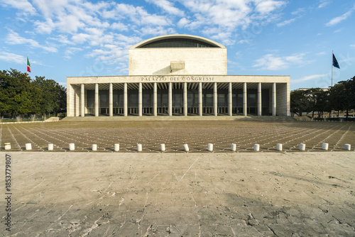 View of the Palazzo dei Congressi, a stark white modernist building, stands imposingly against a backdrop of blue skies and fluffy clouds, Rome, Lazio, Italy.