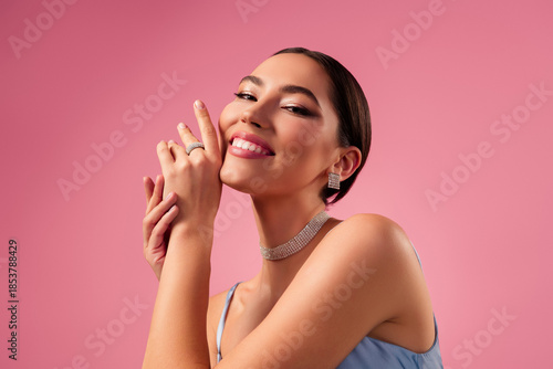 Stunning young woman in elegant dress and jewelry against pink background, showcasing beauty and glamour