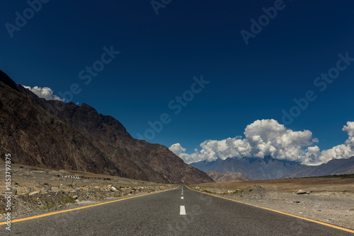 View of an endless asphalt ribbon slicing through the stark, rocky terrain beneath an azure sky dotted with fluffy clouds, Jaglot, Gilgit Baltistan, Pakistan.