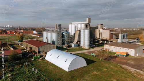 Aerial view of a grain processing plant gleams with metallic silos against a backdrop of industrial buildings and autumn-tinted trees, Sremska Mitrovica, Vojvodina, Serbia.