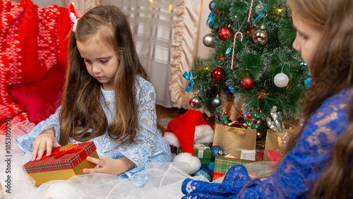 A beautiful little girls in a fluffy festive dress gets surprised with a gift in a Christmas setting. Children's Christmas and New Year.