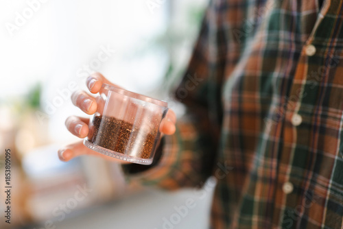 A person in a plaid shirt carefully holds a clear container filled with dark, finely ground material. The individual is examining the contents, possibly for a recipe or a planting project.