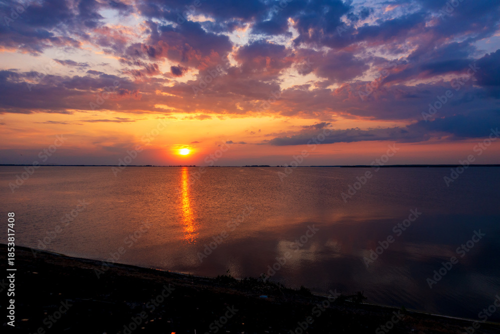 Fototapeta premium Vibrant sunset over a calm lake with golden sun reflection. Dramatic clouds in fiery orange, purple, and blue hues create a picturesque, serene natural landscape.