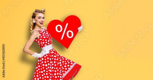 Expressive smile pinup beautiful cute woman point red heart paper banner with percentage percent % discount sign. Pin up girl. Retro concept. Isolate orange yellow background. Valentine or like symbol