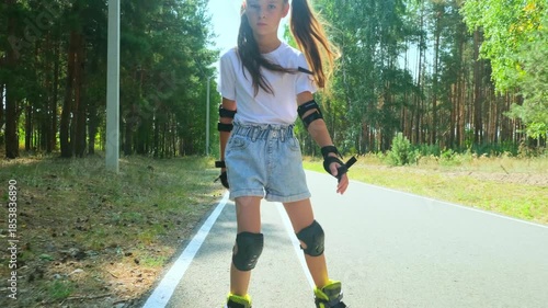 Child girl in casual clothes roller skating on road in forest on sunny summer day.