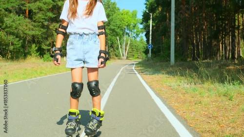 Child girl in casual clothes roller skating on road in forest on sunny summer day.