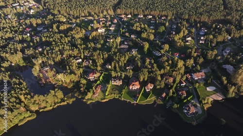 Aerial sweep of lakeside enclave with docks, beach lots, and piers. Drone moves over rooftops and curved shorelines in warm late afternoon light with long tree shadows.