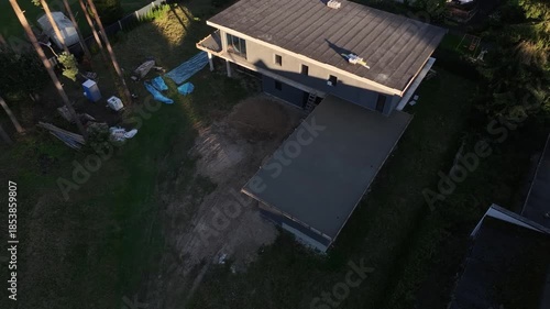 Aerial drone flyover of unfinished flat roof house and pine trees. Title in description. Late afternoon light, slow motion reveals roof details, tarps, slabs, and materials.
