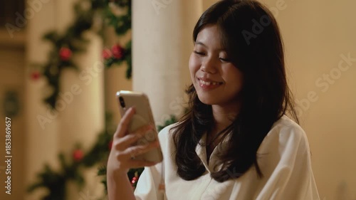 Positive smiling young asian woman is texting someone on mobile phone while at home against the background of a Christmas tree.