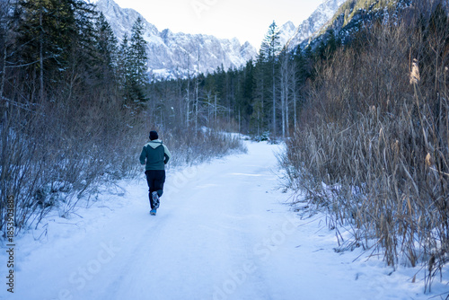 Person running alone on a snow-covered path surrounded by winter forest and mountains