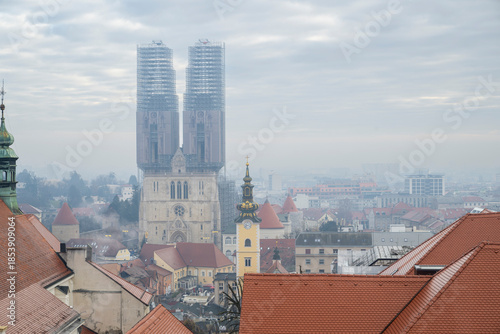 City skyline featuring historic church towers under scaffolding and surrounding red rooftops on a cloudy day