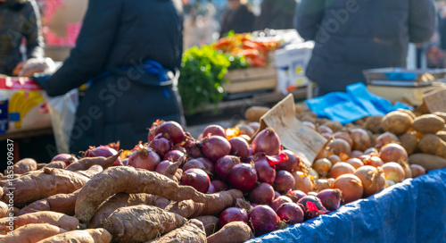 Fresh vegetables including onions and yams displayed at outdoor market with shoppers browsing