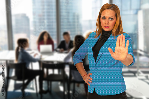 A woman in a blue jacket is standing in front of a group of people in a conference room. She is holding her hand up in a stop sign gesture