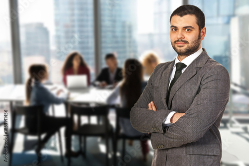 A man in a suit stands in front of a group of people in a conference room. He is wearing a tie and has his arms crossed. The scene suggests a professional setting