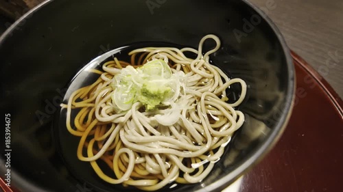 A bowl of clear soba noodles in a black bowl, topped with green onions and wasabi.