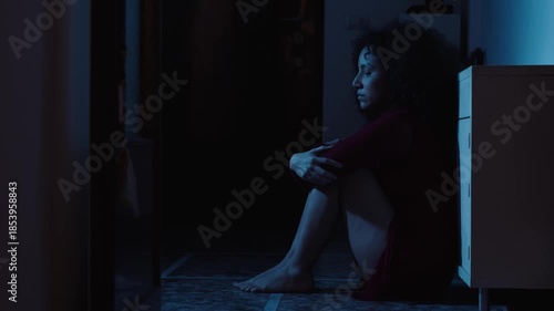 A young Latina woman sits on the floor in a dark room, shown in profile. Her posture and expression reveal deep heartbreak, emotional pain, and loneliness after lost love.