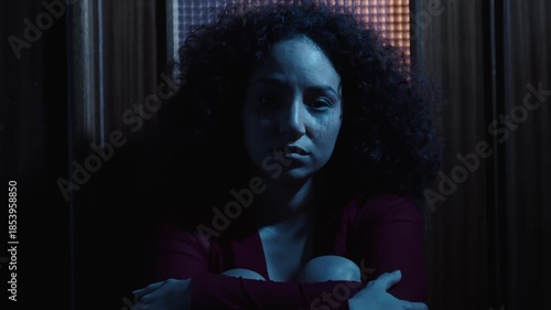 In a dark home environment, a heartbroken young Latina woman sits on the floor and looks directly into the camera, expressing pain, vulnerability, and emotional exhaustion.